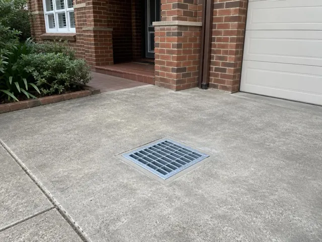 Square outdoor drain grate set in a concrete driveway beside a brick house in Burwood