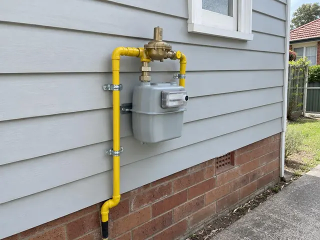 Exterior gas meter with new yellow pipework on a weatherboard wall and brick base in Homebush